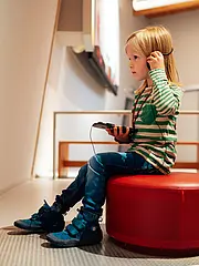 Bild - Ein Kind mit langen blonden Haaren sitzt auf einem roten Hocker und hört über Kopfhörer einem Audiobeitrag zu. Es trägt ein gestreiftes Oberteil und Jeans. Im Hintergrund ist eine Wand mit Ausstellungsobjekten zu sehen.