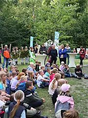 Bild - Eine Gruppe von Kindern sitzt auf dem Boden und hört zwei Rednern zu, die vor ihnen stehen. Im Hintergrund sind Erwachsene versammelt, einige stehen, andere sitzen. Bäume und festliche Dekorationen umgeben den Veranstaltungsort im Freien.