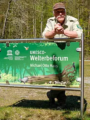 Bild - Ein Mann in Naturbekleidung steht hinter einem Schild, das "UNESCO-Welterbe­forum" und "Michael Otto Haus" anzeigt. Das Schild ist grün gestaltet und zeigt einige Illustrationen von Pflanzen und Tieren. Im Hintergrund ist eine waldreiche Umgebung sichtbar.