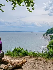 Bild - Zwei Personen sitzen auf einem Baumstamm und blicken auf das Meer. Im Hintergrund sind hohe, bewaldete Klippen und ein bewölkter Himmel zu sehen. Die Szenerie vermittelt Ruhe und Naturverbundenheit.