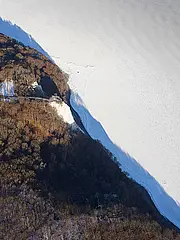 Bild - Luftaufnahme einer bewaldeten Küste, wo Bäume auf einen flachen, gefrorenen Gewässerbereich treffen. Die Landschaft zeigt den Kontrast zwischen den grünen Bäumen und der weißen Eisfläche. In der Nähe der Küste sind einige Gebäude sichtbar, umgeben von der Natur.