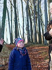 Bild - Drei Personen stehen in einem Wald. Ein Junge spricht gestikuliert, während ein Mädchen aufmerksam zuhört. Ein älterer Mann mit einem Stock lächelt und scheint zu antworten. Es ist ein sonniger Tag und die Umgebung ist von Bäumen und herbstlichem Laub geprägt.
