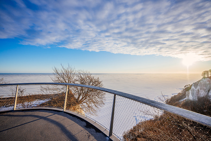 Eine Aussicht auf eine weite, gefrorene Landschaft mit einem klaren Himmel. Die Sonne strahlt am Horizont, während der Blick von einer metallenen Brüstung aus über die schneebedeckte Fläche führt. In der Ferne sind sanfte Hügel und Bäume sichtbar.