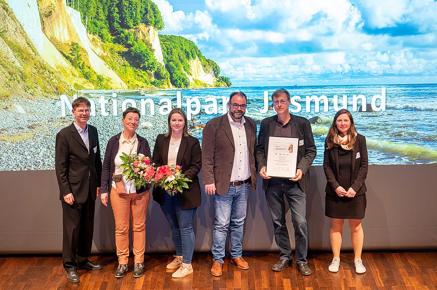Eine Gruppe von fünf Personen steht vor einer großflächigen Hintergrundprojektion des Nationalparks Jasmund. Sie halten Blumen und eine Urkunde. Die Stimmung wirkt feierlich und freundlich.