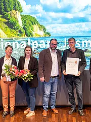 Eine Gruppe von fünf Personen steht vor einer großflächigen Hintergrundprojektion des Nationalparks Jasmund. Sie halten Blumen und eine Urkunde. Die Stimmung wirkt feierlich und freundlich.