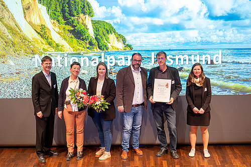 Eine Gruppe von fünf Personen steht vor einer großflächigen Hintergrundprojektion des Nationalparks Jasmund. Sie halten Blumen und eine Urkunde. Die Stimmung wirkt feierlich und freundlich.
