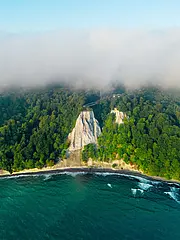Bild - Eine Luftaufnahme zeigt einen Küstenabschnitt mit steilen, weißen Klippen, die von üppigem Grün umgeben sind. Im Hintergrund sind Nebel und Wälder zu sehen, während das Wasser an die Küste brandet.