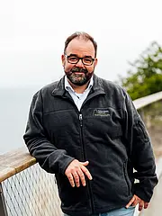 Bild - Ein Mann steht auf einem Aussichtssteg mit Blick auf das Wasser. Er trägt eine dunkle Jacke mit einem Logo und hat eine Brille. Der Hintergrund ist unscharf, mit etwas Grün und einem grauen Himmel sichtbar.