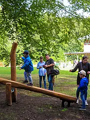 Bild - Ein Junge spielt auf einer Wippe im Freien, während mehrere Kinder und Erwachsene zuschauen. Umgeben von Bäumen und einer Wiese, ist der Hintergrund verschwommen und zeigt ein Gebäude, möglicherweise ein Restaurant oder einen Spielplatz.