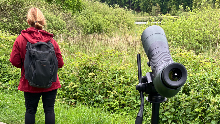Eine Person mit einem Rucksack steht im Hintergrund in einer grünen Landschaft. Im Vordergrund steht ein Stativ mit einem Fernglas, das auf die Umgebung ausgerichtet ist, um die Natur zu beobachten.