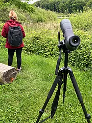 Eine Person mit einem Rucksack steht im Hintergrund in einer grünen Landschaft. Im Vordergrund steht ein Stativ mit einem Fernglas, das auf die Umgebung ausgerichtet ist, um die Natur zu beobachten.