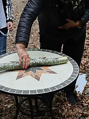 Bild - Eine Person rollt einen grünen Gegenstand auf einem runden Tisch mit einem geometrischen Muster aus Stein. Um den Tisch stehen weitere Personen, die gespannt zuschauen. Der Boden ist mit trockenen Blättern bedeckt.