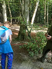 Bild - Eine Gruppe von Kindern steht im Wald und hört einem Mann zu, der ihnen etwas erklärt. Er trägt eine grüne Jacke und hält ein Objekt in der Hand. Die Umgebung besteht aus Bäumen und blättrigem Boden. Die Kinder wirken neugierig und aufmerksam.