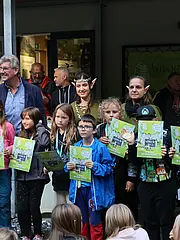 Bild - Eine Gruppe von Kindern hält stolz Urkunden in den Händen. Sie stehen in einem Außenbereich, umgeben von mehreren Erwachsenen. Einige Kinder tragen Medaillen oder Bänder um den Hals. Im Hintergrund sind Bäume und eine Willkommensschild sichtbar. Die Atmosphäre ist feierlich und fröhlich.