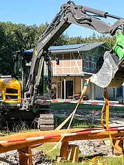 Bild - Ein gelber Bagger steht auf einer Baustelle umgeben von Absperrbändern. Im Hintergrund ist ein modernes Gebäude mit Holzverkleidung und Fenstern zu sehen. Der Bagger hat eine große Schaufel, die nach unten zeigt, während einige Personen auf dem Weg im Hintergrund vorbeigehen.