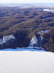 Bild - Luftaufnahme einer Winterlandschaft mit schneebedecktem Boden und schroffen Klippen. Die Küste wird von Wäldern umgeben, und im Hintergrund sind sanfte Hügel sowie ein weiter Wasserbereich zu sehen. Die gesamte Szene vermittelt ein Gefühl von Ruhe und unberührter Natur.