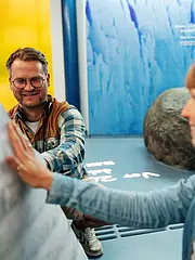 Bild - Zwei Personen interagieren in einem Ausstellungsraum. Eine Frau berührt eine große, strukturierte Oberfläche, während ein Mann lächelnd auf sie schaut. Beide haben Kopfhörer auf und scheinen interessiert an der Ausstellung zu sein. Bunte Hintergründe und informative Elemente sind sichtbar.