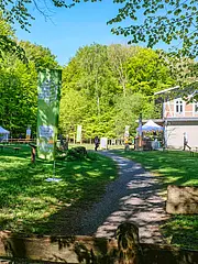 Bild - Ein schmaler Weg führt durch eine grüne Wiese, umgeben von Bäumen. Links steht ein Holzzaun, rechts ein modernes Gebäude mit Fenstern. Im Hintergrund sind Tische, ein Zelt und einige Personen zu sehen, die die Umgebung genießen. Klarer blauer Himmel über der Szenerie.