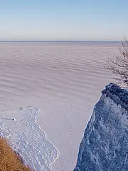 Bild - Eine schneebedeckte Klippe ragt über eine gefrorene Wasserfläche. Die Landschaft ist weitläufig, mit sanften Wellen im Eis und wenigen kahlen Bäumen am Rand der Klippe. Der Himmel ist klar und hell, was eine ruhige und frostige Stimmung vermittelt.