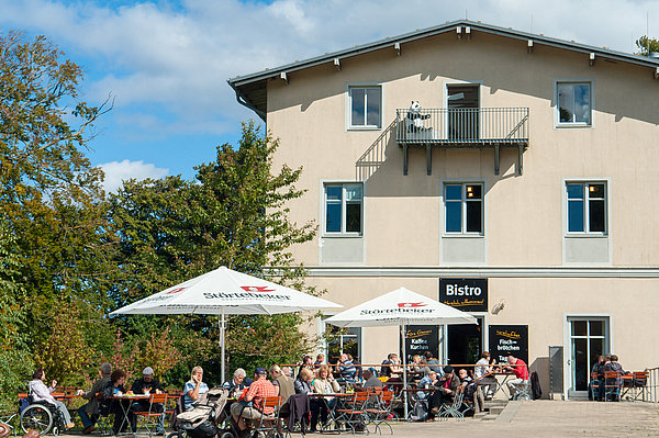 Eine Gruppe von Menschen sitzt an Tischen vor einem Bistro unter Sonnenschirmen. Im Hintergrund steht ein helles Gebäude mit Fenstern und einem Balkon. Bäume umgeben den Bereich, und der Himmel ist teils bewölkt.