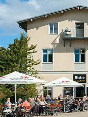 Bild - Eine Gruppe von Menschen sitzt an Tischen vor einem Bistro unter Sonnenschirmen. Im Hintergrund steht ein helles Gebäude mit Fenstern und einem Balkon. Bäume umgeben den Bereich, und der Himmel ist teils bewölkt.