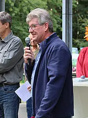 Bild - Mehrere Personen stehen auf einer Veranstaltung. Ein Mann mit Mikrofon spricht, während ein anderer eine Box in der Hand hält. Im Hintergrund sind Menschen und grüne Landschaft sichtbar. Eine Frau trägt einen Kopfschmuck aus bunten Blättern.