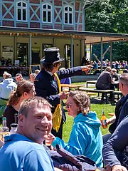 Bild - Eine Gruppe von Menschen sitzt an Tischen im Freien, umgeben von Bäumen. Jemand im Anzug und Zylinder zeigt mit dem Finger auf etwas, während andere Gäste entspannt Getränke genießen und sich unterhalten. Im Hintergrund ist ein Gebäude mit Fenstern und einem Banner sichtbar.