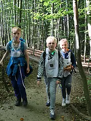 Bild - Drei Kinder wandern auf einem schmalen Waldweg. Sie tragen Rucksäcke und sehen fröhlich aus. Bäume umgeben den Weg, und es ist tagsüber mit natürlichem Licht erhellt. Der Boden ist mit Laub und Erde bedeckt.