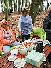 Bild - Eine Gruppe von Frauen und einem Jungen sitzt in einem Wald. Sie tragen fröhliche Hüte mit Blumen und Blättern. Vor ihnen steht ein Tisch mit Teetassen, einem Kaffeekrug und einer dekorativen Schachtel. Die Atmosphäre ist freundlich und einladend.