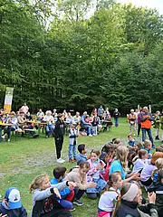 Bild - Eine große Gruppe von Menschen, darunter viele Kinder, sitzt auf dem Boden und schaut aufmerksam nach vorne. Einige Erwachsene stehen oder sitzen an Tischen im Hintergrund, während Bäume die Umgebung umgeben. Es findet offensichtlich eine Veranstaltung oder Aufführung im Freien statt.