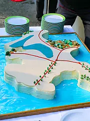 Bild - Ein großer, dekorierter Kuchen in Form einer Karte zeigt unterschiedliche Landschaftselemente, darunter Wasserflächen und grüne Vegetation. Der Kuchen ist auf einem Tisch platziert und umgeben von grünen Tellern. Das Gesamtbild vermittelt eine festliche Atmosphäre.