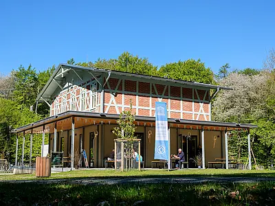 Ein modernes Gebäude im Freien, umgeben von Bäumen. Vor dem Gebäude befinden sich einige Tische und Bänke, an denen Menschen sitzen. Eine blaue Fahne mit einem Logo ist sichtbar. Der Himmel ist klar und sonnig, was eine einladende Atmosphäre schafft.