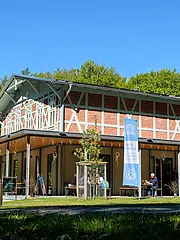 Bild - Ein modernes Gebäude im Freien, umgeben von Bäumen. Vor dem Gebäude befinden sich einige Tische und Bänke, an denen Menschen sitzen. Eine blaue Fahne mit einem Logo ist sichtbar. Der Himmel ist klar und sonnig, was eine einladende Atmosphäre schafft.