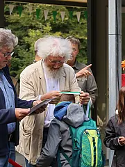 Bild - Eine Gruppe von Erwachsenen und Kindern versammelt sich im Freien. Ein älterer Mann mit weißem Haar lächelt, während er einem Jungen mit einem Rucksack etwas zeigt. Im Hintergrund schaut eine Frau mit einer bunten Kopfbedeckung aufmerksam zu. Die Atmosphäre wirkt freundlich und informell.