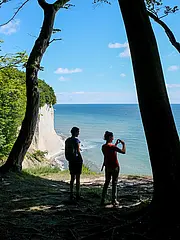Bild - Zwei Personen stehen am Rande einer Klippe mit Blick auf das Meer. Die Landschaft ist von Bäumen umgeben, und einer der Personen macht ein Foto. Das Wetter ist klar mit blauen Himmel und wenigen Wolken.