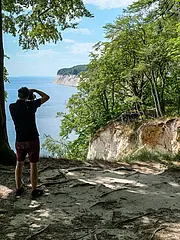 Bild - Ein Mann steht an einem felsigen Aussichtspunkt im Wald und fotografiert die Küste. Hinter ihm ist ein weiterer Mann mit einem Rucksack zu sehen. Der Blick geht über das Wasser und die Felsen, umgeben von Bäumen und hellem Himmel.