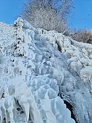 Bild - Eine vereiste Steigung ist zu sehen, bedeckt mit gefrorenem Wasser und Schnee. Die Struktur hat eine unregelmäßige, wellenförmige Oberfläche. Im Hintergrund sind kahle Bäume unter einem klaren blauen Himmel angedeutet.