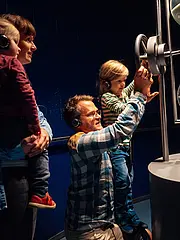 Bild - Eine Familie mit zwei Kindern betrachtet eine große Sanduhr in einem Lernbereich. Die Erwachsenen tragen Kopfhörer und scheinen interessiert. Ein Kind steht neben der Sanduhr und untersucht sie, während das andere auf dem Arm eines Elternteils sitzt.