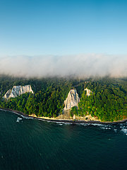 Luftaufnahme einer grünen, bewaldeten Insel, die von sanften Klippen umgeben ist. Der Himmel ist leicht bewölkt, der Ozean zeigt verschiedene Blautöne. Die Klippen sind hell und auffällig, während das Ufer sanft ins Wasser übergeht.