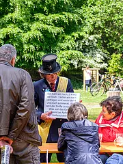 Bild - Eine Gruppe von Menschen hört einem Mann im Zylinder und Anzug zu, der ein Schild mit Text hält. Der Mann liest humorvoll über verschiedene Geschmäcker von Gerichten. Im Hintergrund sind Bäume und weitere Personen bei einer Veranstaltung zu sehen.