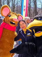 Bild - Drei Personen posieren fröhlich in einem Park. Eine Frau lächelt neben einem großen, verkleideten Maulwurf mit rotem Schal. Eine weitere Frau steht mit einem schwarzen Vogel-Maskottchen. Im Hintergrund sind verschwommene Menschen und Bäume sichtbar.
