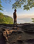 Eine Person steht auf einem Aussichtspunkt, umgeben von Bäumen, und schaut auf das Meer. Die Sonne scheint, und der Himmel ist teilweise bewölkt. Der Boden zeigt natürliche Unebenheiten und Erde, wodurch die Szenerie eine ruhige, naturnahe Atmosphäre ausstrahlt.