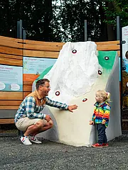 Bild - Ein Erwachsener und ein Kind betrachten gemeinsam eine interaktive Tafel, während eine weitere Person daneben steht. Die Tafel zeigt eine Berglandschaft mit farbigen Markierungen. Hintergrund: Baumgruppe und Natur.