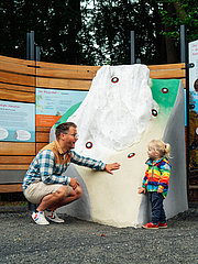 Ein Erwachsener und ein Kind betrachten gemeinsam eine interaktive Tafel, während eine weitere Person daneben steht. Die Tafel zeigt eine Berglandschaft mit farbigen Markierungen. Hintergrund: Baumgruppe und Natur.