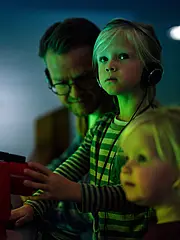 Bild - Eine Gruppe von Erwachsenen und Kindern steht in einem dunklen Raum und betrachtet ein Exponat. Die Kinder tragen Kopfhörer und schauen interessiert. Ein Junge hält ein Fernrohr, während eine Frau und ein Mädchen ebenfalls aufmerksam nach vorne blicken.