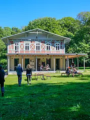 Bild - Eine Gruppe von Personen nähert sich einem großen, hellen Gebäude mit einem Holzdach und Fenstern im oberen Bereich. Vor dem Gebäude sind Tische im Freien, an denen Menschen sitzen und entspannen. Umgeben von Bäumen und grünem Rasen ist der Himmel klar und blau.