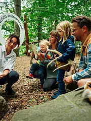 Bild - Eine Gruppe von Erwachsenen und Kindern untersucht gemeinsam etwas im Freien. Eine Person zeigt ein Buch, während die anderen interessiert zuschauen. Im Hintergrund ist ein Schild mit der Aufschrift „UNESCO-Weltnaturerbe“ zu sehen, umgeben von Bäumen und Steinen.