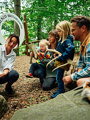 Eine Gruppe von Erwachsenen und Kindern untersucht gemeinsam etwas im Freien. Eine Person zeigt ein Buch, während die anderen interessiert zuschauen. Im Hintergrund ist ein Schild mit der Aufschrift „UNESCO-Weltnaturerbe“ zu sehen, umgeben von Bäumen und Steinen.