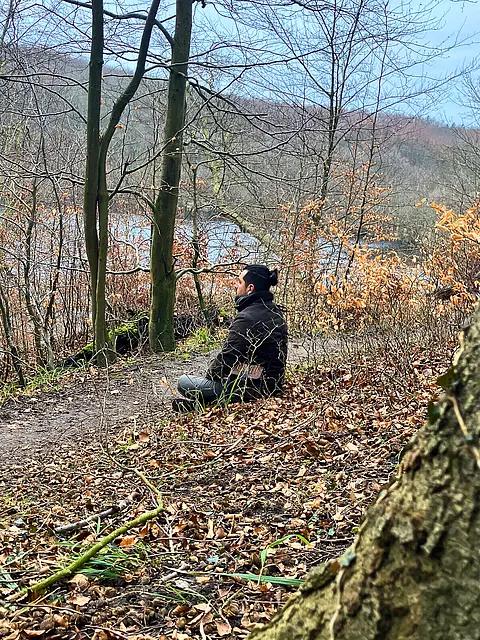Ein Mann sitzt am Waldrand auf einem Weg und blickt auf einen stillen See, umgeben von kahlen Bäumen und vereinzeltem, braunem Laub. Die Umgebung vermittelt eine ruhige, natürliche Atmosphäre.