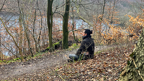 Ein Mann sitzt am Waldrand auf einem Weg und blickt auf einen stillen See, umgeben von kahlen Bäumen und vereinzeltem, braunem Laub. Die Umgebung vermittelt eine ruhige, natürliche Atmosphäre.